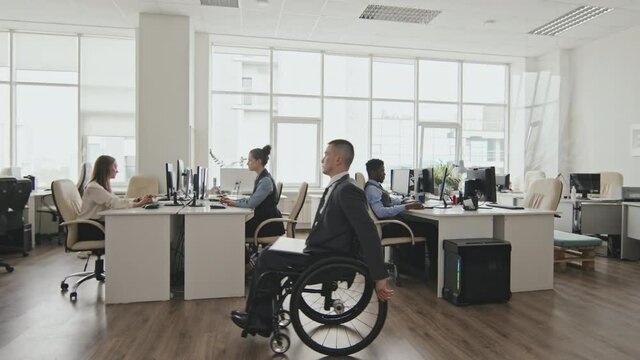 Wide shot of disabled businessman in wheelchair riding through open-plan office. Caucasian businesswomen and African-American businessman working on computers