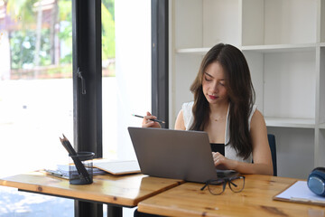 Young female freelancer holding pencil and working with laptop computer at coffee shop.
