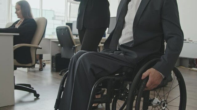 Tracking mid-section shot of unrecognizable businesswoman walking beside disabled businessman riding wheelchair in office and showing him something on tablet