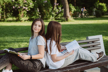 Two student girls are looking at an open book on a bench in the park. Distance education, preparation for exams. Soft selective focus.