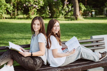 Two student girls are looking at an open book on a bench in the park. Distance education, preparation for exams. Soft selective focus.