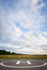 A closeup of an asphalt-covered helipad with a special symbol in the center for helicopter landing, against the backdrop of a green field and a cloudy evening sky.