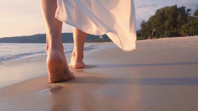 4K Slow motion woman feet walking barefoot on the beach at golden sunset time. leaving footprints in sand. Female tourist on summer vacation holiday relax at tropical beach Phuket, Thailand.