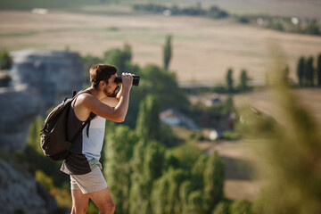 Guy looking at binoculars in hill. man in t-shirt with backpack.