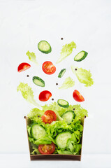 Fresh summer salad of raw vegetable with flying ingredients - tomatoes, cucumber, green salad above craft box for take away food in soft light interior, vertical.