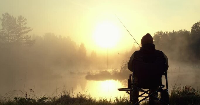 A man in warm clothes sits on a chair by the lake in the early morning and goes fishing. The bright sun illuminates the fisherman, so only his silhouette is visible. There is fog over the reservoir