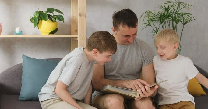 Father with two young sons watches the family photo album sitting on a couch in the living room. Family values. Front view of natural daylight