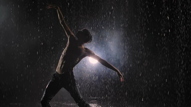 Emotional performance of modern contemporary choreography in rain against bright spotlight. Puffs of smoke hide silhouette of male dancer with naked torso among drops of water. Slow motion.