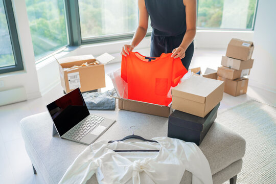 Selling new or used clothes online with internet app on laptop computer screen. Woman packing fashion clothing in mailing box for shipping orders from home. Small business.