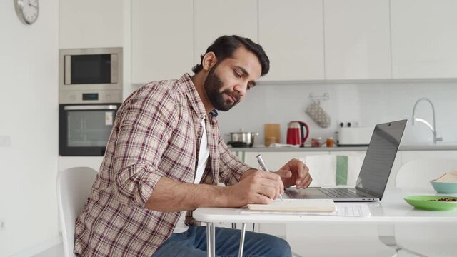 Young Indian Hispanic businessman using computer remote distance working sitting at kitchen table writing notes. Latin student studying online, having virtual digital training on laptop at home office