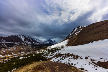 Fototapeta na wymiar A spring storm in the distance is creating dramatic clouds over this glacial valley in the Chugach Mountains, Alaska. Snow-covered mountains, taiga forests, and alpine tundra surround the valley.