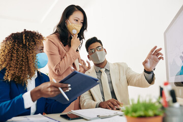 Business team in protective masks looking at chart on computer screen and discussing sales growth - Powered by Adobe