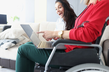 Disabled woman in wheelchair with a laptop on her knees shows her joyful girlfriend her...
