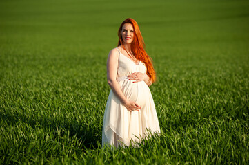 Pregnant woman on the summer green field wearing long white dress