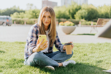 Young caucasian Woman having a video call chat with cellphone, sitting on the grass outside in park. Happy and smiling girl relax outdoors. Using mobile phone. Distance learning online education.