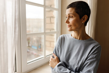 Portrait of sad middle aged female with short hairstyle posing by window looking outside with bored...