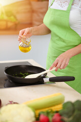 An attractive young dark-haired woman frying meal at worm pan by new keto recipe while standing and smiling in sunny kitchen. Cooking and householding concepts