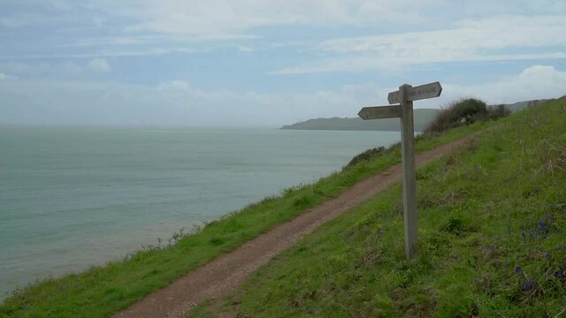 Footage panning across seascape scenery with South West devon coastal path sign in foreground on green hills with path way leading into distance. Start point lighthouse and cliffs in the background.