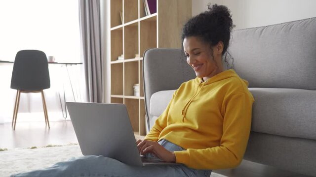 Young African American adult student sitting on floor typing on laptop computer having virtual education class, e learning webinar, shopping online or searching new job on device at home office.