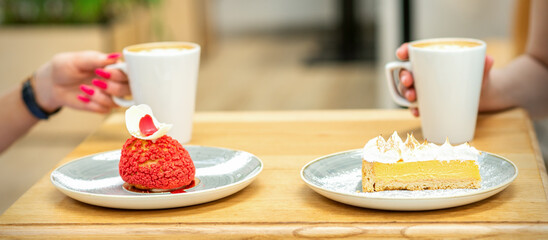 Two young girlfriends drinking coffee with pieces of cake sitting at the table in a cafe outdoors