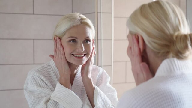 Attractive beautiful mid age adult 50 years old blonde woman standing in bathroom touching face with hands, smiling to reflection in mirror doing daily morning beauty routine. Older skin care concept.