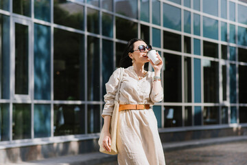 Brunette young woman with sunglasses and bag holding coffee walking in the city. Lifestyle portrait of woman