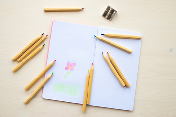 Open white notebook, with colored crayons beside it, penknife and drawing of a colored flower. 