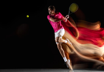 Young caucasian male tennis player playing tennis in mixed light on dark background.