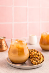 Cold iced coffee with milk in a glass, served with oatmeal cookies. A cezve and white milk jug. Warm light and bright vertical image, pink tile backdrop