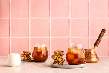 Cold iced coffee in a glass, served with oatmeal cookies. A cezve and white milk jug. Warm light and bright image, pink tile backdrop