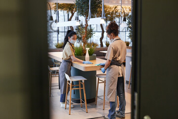 Full length portrait of two female workers wearing masks while cleaning tables in modern cafe interior, covid safety concept