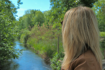 Beautiful blonde woman with long blonde hair, in profile, at the water's edge in nature. Lake and vegetation blurred voluntarily in the background.