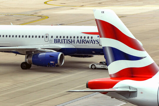 London, England - April 2018: Tail Fin Of A British Airways Jet With A Jet Taxiing In The Background