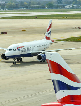 London, England - April 2018: Tail Fin Of A British Airways Jet With A Jet Taxiing In The Background