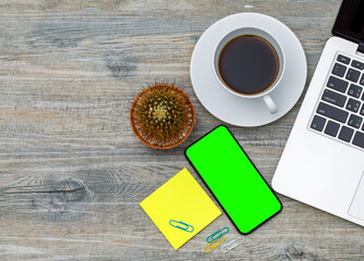 Topview hign angle shot of laptop notebook computer with green blank screen smartphone and yellow paper notepad with coffee cup on wooden backgroundd and copy space