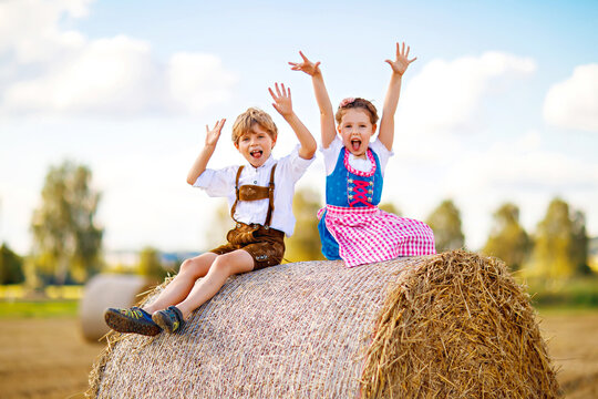 Two kids in traditional Bavarian costumes in wheat field. German children sitting on hay bale during Oktoberfest. Boy and girl play at hay bales during summer harvest time in Germany. Best friends.