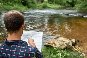 The man in a plaid shirt standing on the bank of the river points a finger to a place on the map
