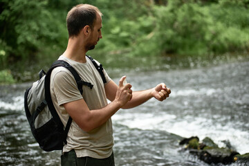 Side view of a hiker in a shirt with a backpack near the river spraying insect repellent on skin.
