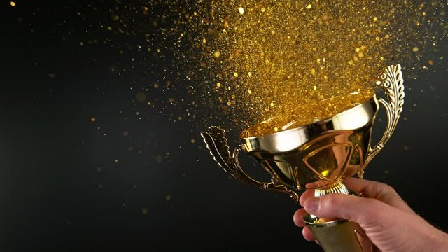 Super Slow Motion. Champion Golden Trophy with Exploding Gold Glitters and Stars against black background. Concept of Success, Victory or Achievement. 1000 fps. Filmed on high Speed Cinematic Camera.