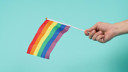Hand is holding a rainbow flag on mint green or tiffany blue background.