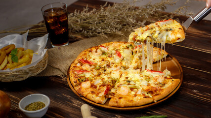 Selective focus and close up on a piece of double cheese homemade seafood pizza on spatula or scoop with blur background of wooden table, sackcloth, ingredients and fried potatoes. Food Concept.
