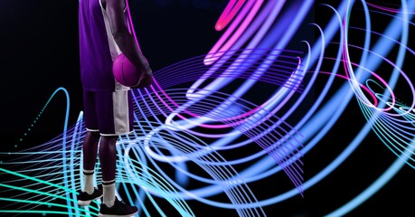 Composition of midsection of basketball player with ball over neon light trails on black background