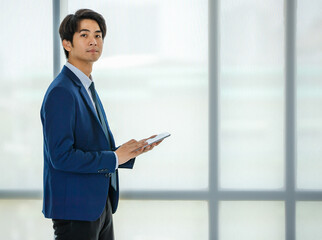 Fototapeta premium 20s young confident attractive Asian businessman wearing formal suit, standing and putting hand in pocket pants, holding tablet and looking at the camera. Showing modernity, startup, and technology.