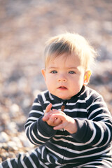 Serious baby in striped overalls sitting on a pebble beach holding folded palms in front of him