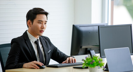 Young Asian businessman in a formal black suit looks elegant. Sitting in front of the computer in the office, white tone. With skill and determination. He deserves to be the leader of the company.