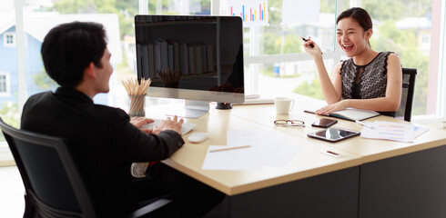 Banner and Selective focus on Asian cheerful business working woman wearing casual shirt, laughing with happiness, sitting at desk in indoor office, holding pen with blur foreground of male colleague.