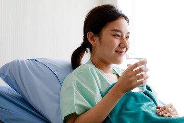 An asian young beautiful female patient lying on bed in the hospital, holding a glass and drinking...