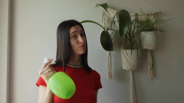 A young Woman looks at the camera smiles and talks about taking care of plants. Beautiful woman holding spray bottle near plants. Indoor plants in macrame plant hanger hang on the wall.