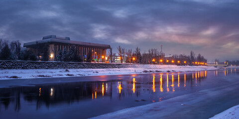 uzhhorod, ukraine - 26 DEC 2016: winter cityscape at dawn. beautiful scenery on the river uzh. city lights reflecting in the water. snow on the embankment