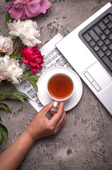 Coffe, peonies and laptop on grey vintage background. Women hand holds cup of coffee.